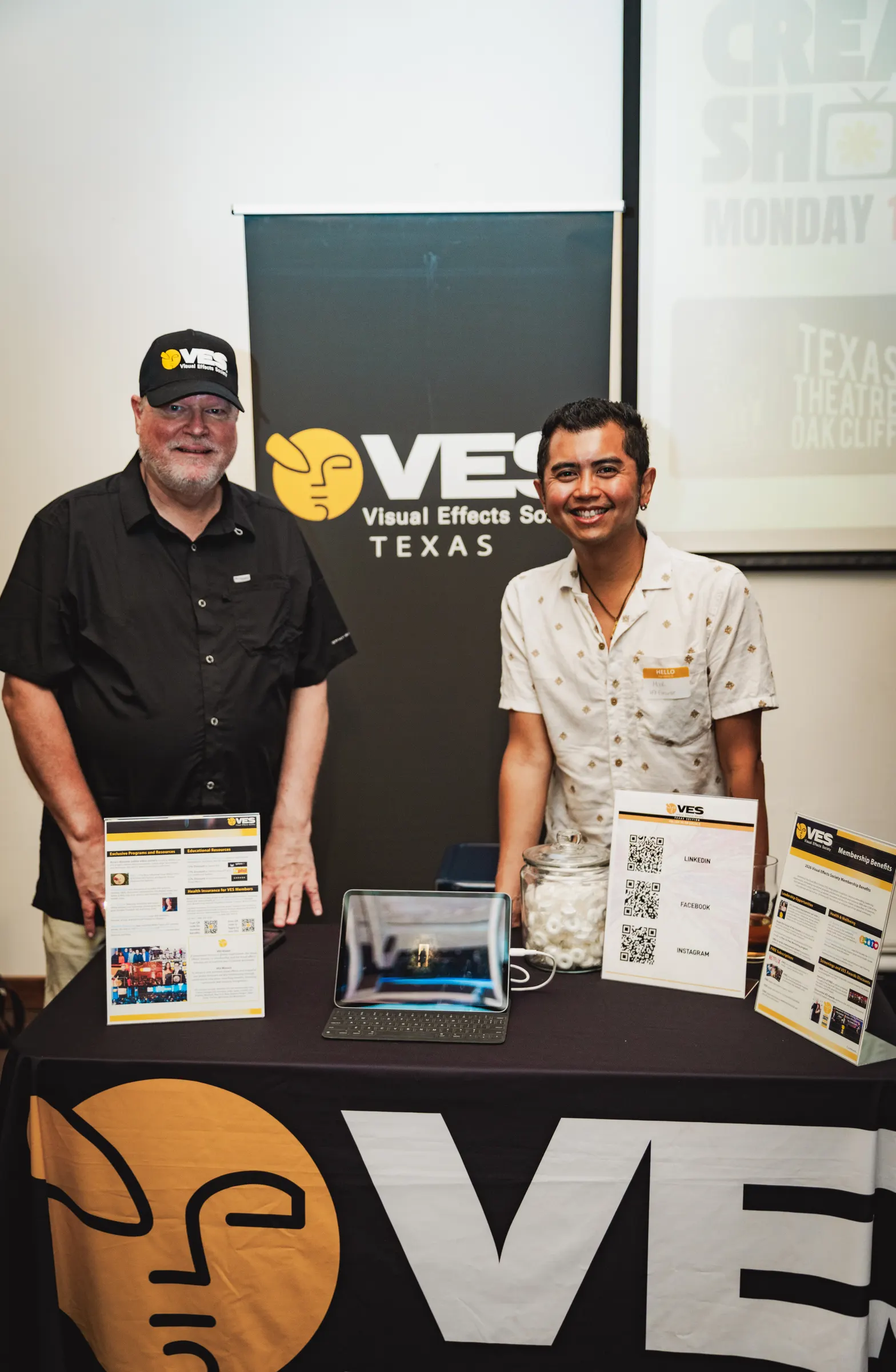 Two exhibitors behind the VES Texas table, with flyers and sign-up QR codes